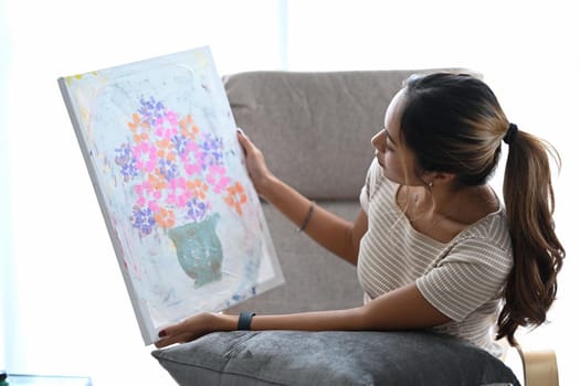 Pleased young woman sitting on armchair in living room and looking at her artwork with admiration.