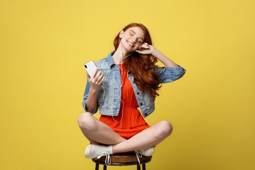 Lifestyle, Music, Technology concept: Young beautiful caucasian woman listening music with headphones and smart phone hand hold, dancing, eyes closed smiling, Isolated on yellow vivid background