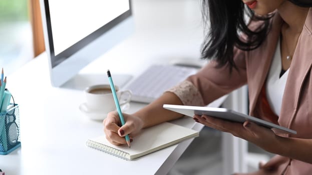 Female office worker using digital tablet and making notes on notebook.