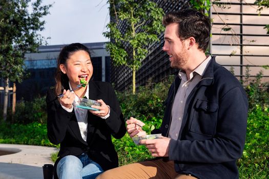 asian woman laughing and eating a salad with her caucasian partner in a park next to the office, concept of healthy fast food at work