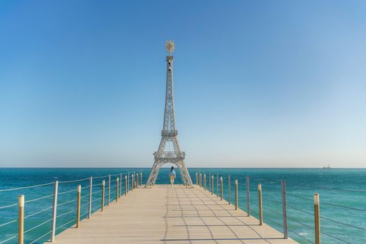 Large model of the Eiffel Tower on the beach. A woman walks along the pier towards the tower, wearing a blue jacket and white jeans