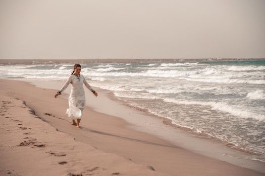 Model in boho style in a white long dress and silver jewelry on the beach. Her hair is braided, and there are many bracelets on her arms