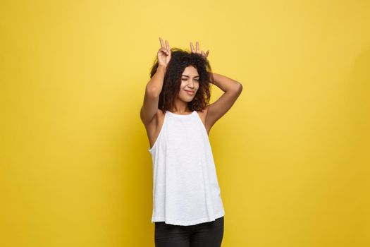 Fun and People Concept - Headshot Portrait of happy Alfo African American woman with freckles smiling and showing rabbit ears with fingers over head . Pastel yellow studio background. Copy Space
