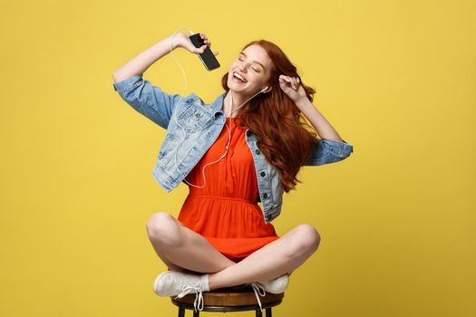 Lifestyle Concept - Happy pretty red hair woman in earphones listening to music and singing while sitting on chair and holding mobile phone. Isolated on bright yellow background.