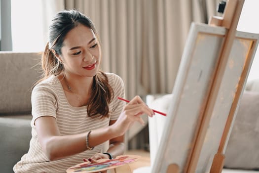 Positive young woman holding paintbrush and palette painting on canvas.