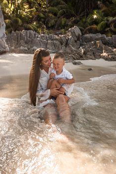 Mother plays with her son in the ocean. Happy smile european family have rest and run on white sand beach. long black chestnut hair mother, blond baby boy. white cotton clothes. boho dress.Thailand.