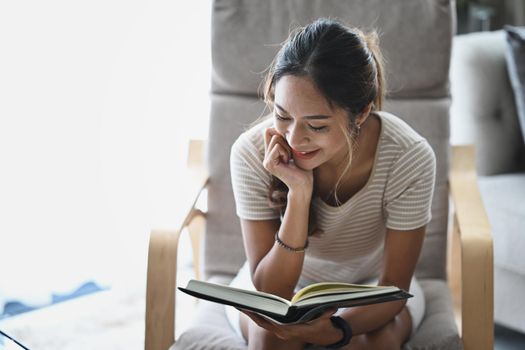 Satisfied young woman reading book on armchair while spending leisure time at home.