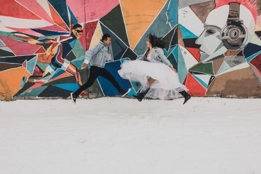 Khmelnytskyi, Ukraine - 27.11.2018: cool young newlyweds in wedding clothes and denim jackets jumping against the background of the wall painted pop art street art in winter in the snow. dynamic and cheerful photo of the bride and groom