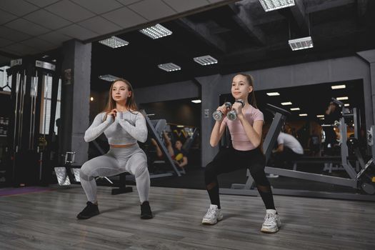 Personal trainer and her teenage female client doing squats with dumbbells together