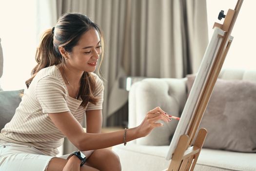 Charming young woman painting picture on canvas with oil paints at home.