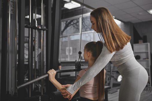 Rear view shot of personal trainer helping teenage girl exercise on lat pull down gym machine