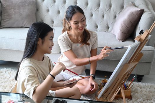 Smiling asian woman and her young sister sitting in front of easel and painting picture on canvas.
