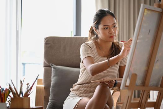 Casual young woman sitting in front of easel and painting picture on canvas.