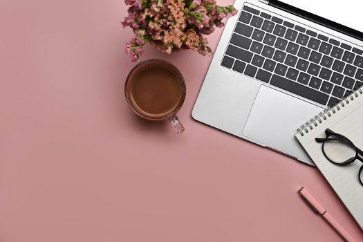 Above view feminine workspace with laptop, notepad, flowers and coffee cup on pink background.