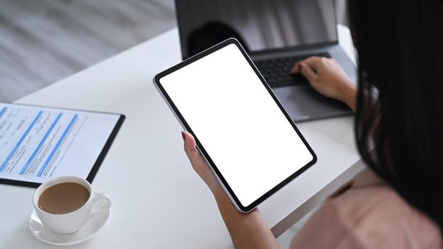 Over shoulder view of businesswoman holding digital tablet and using laptop computer.