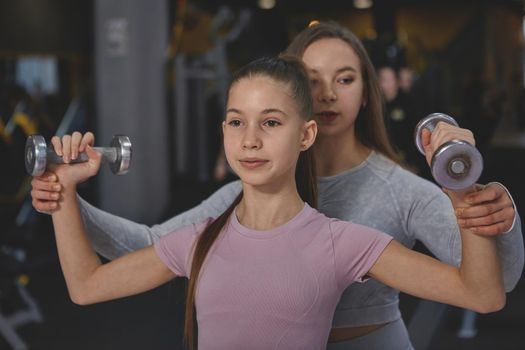 Lovely teenage girl lifting dumbbells, exercising with professional female athlete at the gym
