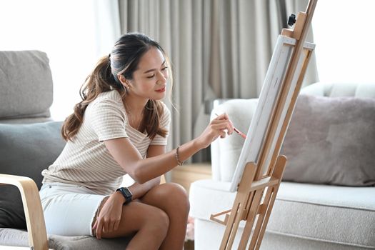 Relaxed young woman sitting in front of easel and painting picture on canvas.