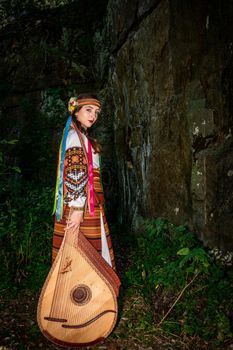 Ukrainian woman in national dress with a musical instrument bandura stands by a rock.