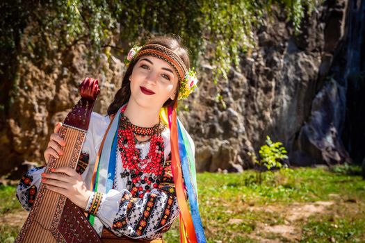 Ukrainian musician in authentic national dress sits by a rock. Ukrainian woman with a bandura musical instrument before performing folk music