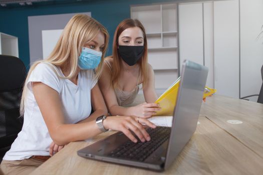 Two young women wearing protective face masks, working on a laptop together at open space office during coronavirus