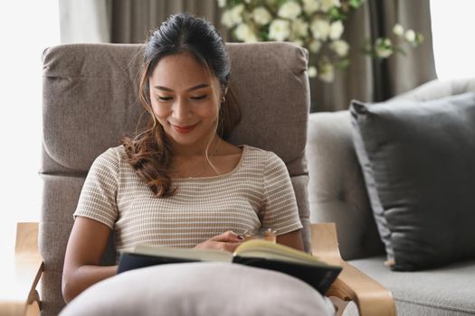 Relaxed young woman sitting on armchair in living room and reading her favorite novel book.