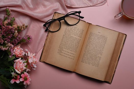 Above view vintage book, eyeglasses coffee cup and spring flowers on pink background.