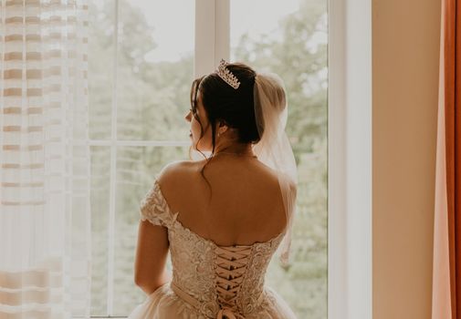 European Caucasian young black-haired woman bride in white wedding dress with long veil and tiara on head. girl stands with her back near window morning gatherings of preparing bride for holiday party