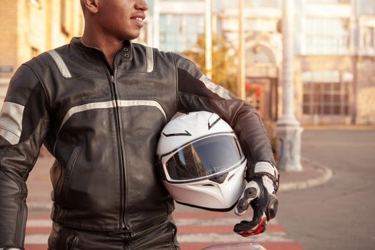 Cropped shot of a young male biker smiling, holding protective helmet, wearing motorbike clothing, copy space. Road trip, protective gear concept