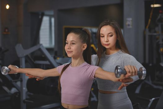 Professional sportswoman supervising teenage girl during gym workout, doing dumbbells exercise