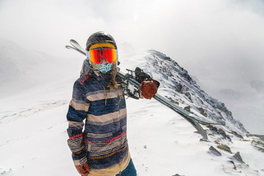 Portrait of a pretty and active woman skier, wearing a mask and holding skis in her hands, active winter holidays. extreme lifestyle concept