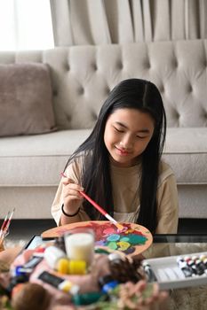 Smiling asian girl holding painting brush and mixing color oil painting on palette.