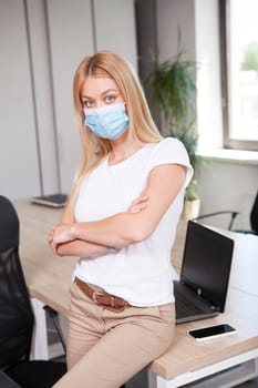 Vertical portrait of a businesswoman posing confidently at her office, wearing medical face mask