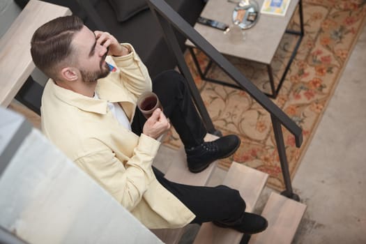 Top view shot of a handsome bearded man talking on the phone, sitting on the staircase at his apartment