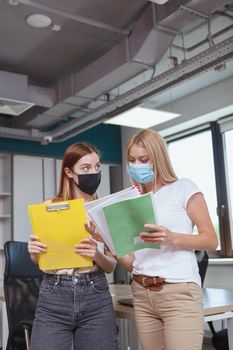 Vertical shot of two female colleagues wearing face masks, working at the office during covid pandemic