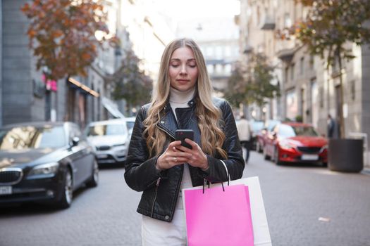 Beautiful stylish woman with shopping bags texting on her smart phone standing on the street