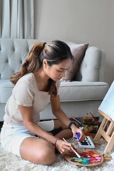 Creative young woman sitting in front of easel and painting picture with water color.