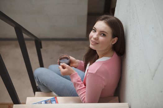 Top view shot of a lovely young woman smiling to the camera while drinking tea at her apartment, sitting on the staircase