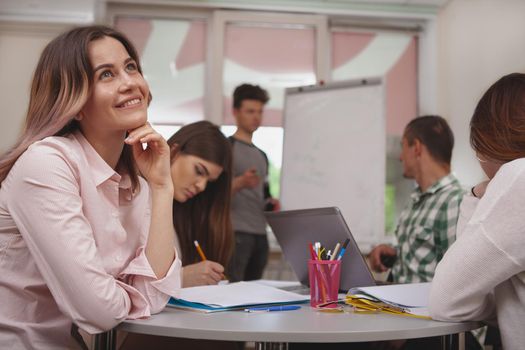 Beautiful happy young female student looking away dreamily, her classmates discussing study project on the background. College students during project presentation at class