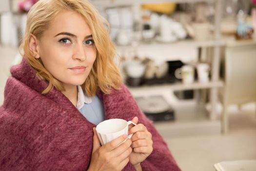 Attractive young woman smiling to the camera holding cup of coffee sitting comfortably wrapped in warm woolen blanket home recreation weekend lifestyle living leisure beauty serenity homeware