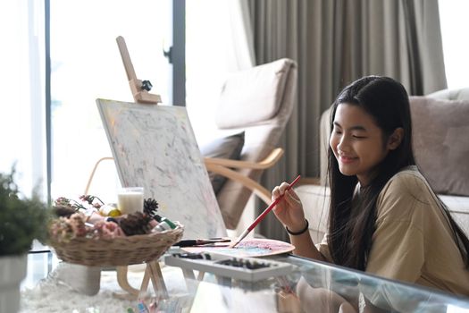 Joyful asian girl sitting in living room and painting picture with watercolor.