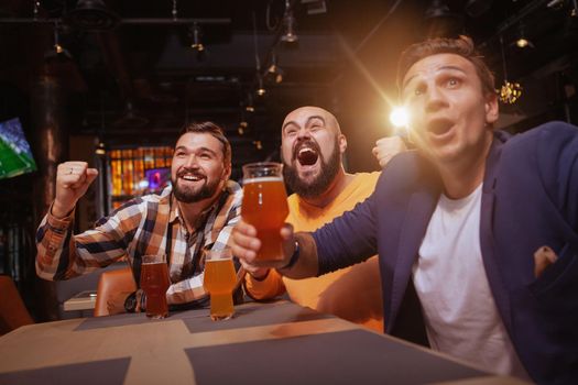 Low angle shot of three excited men screaming happily, celebrating victory of their favourite football team. Soccer fans drinking at beer pub, watching a game