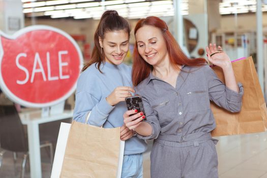 Two cheerful female friends laughing, using smart phone at the shopping mall
