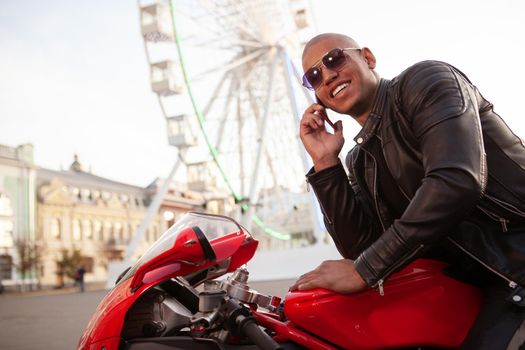 Happ handsome young man laughing, talking on the phone, sitting on a motorcycle. Cheerful African male biker using smart phone while travelling on a motorcycle