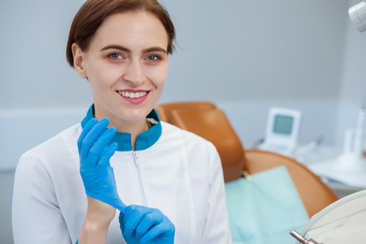 Close up of a beautiful female dentist smiling to the camera, putting on medical rubber gloves, preparing for work at her clinic, copy space. Attractive dentist posing at her office confidently