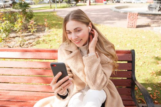 Attractive woman smiling, using earphones with her smart phone, sitting on park bench