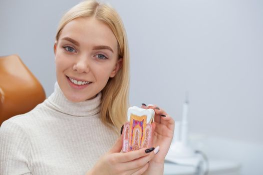 Beautiful young woman smiling to the camera, sitting at dentist office, copy space. Attractive female patient holding tooth mold, posing at dental clinic after oral examination. Health, medicine