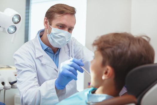 Professional dentistry, healthy teeth concept. Mature male dentist wearing medical mask and gloves, examining teeth of a little boy
