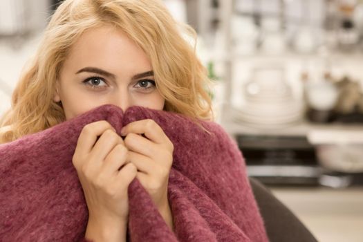 Close up of a happy blonde woman peeking to the camera sitting wrapped in a warm woolen blanket hiding her face playfully relaxing at home copyspace lifestyle living positivity youth homeware comfort