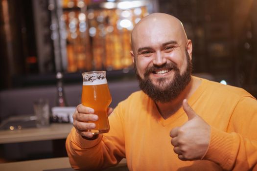 Excited bearded bald man smiling to the camera, showing thumbs up while having a glass of delicious beer at the pub