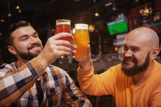 Happy male friends laughing, toasting with their beer mugs. Handsome bearded men clinking beer glasses, celebrating at the pub